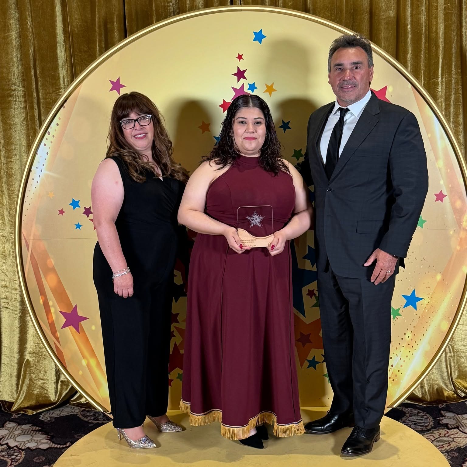 Maribel Rodriguez holding her award on stage at an awards ceremony, standing between two colleagues